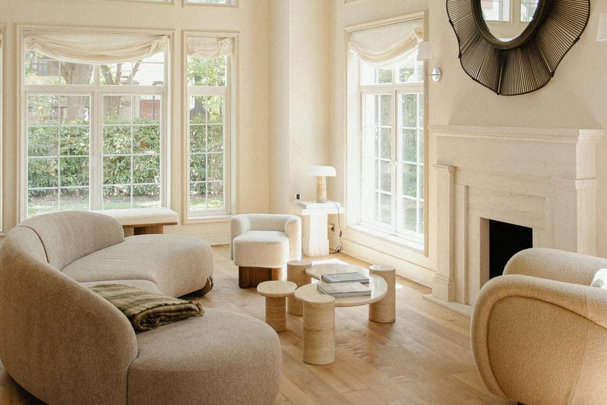 A bright, high-ceilinged living room with cream walls, tall windows, light wood floors, a curved upholstered sofa, matching armchairs, a fireplace, and a decorative sunburst mirror.
