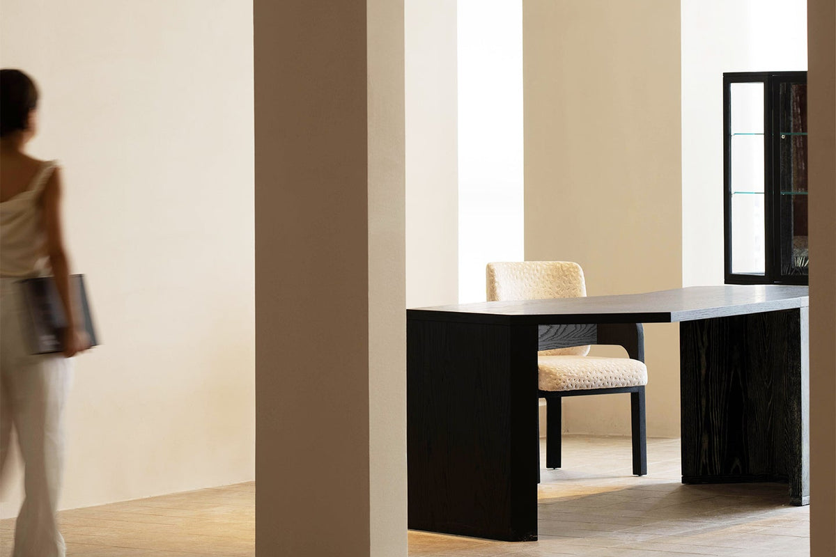 A minimalist interior scene with warm beige walls and arches, featuring a black desk with a cream upholstered chair. A woman in light clothing walks through the left arch, and a glass cabinet is visible in the background on the right.