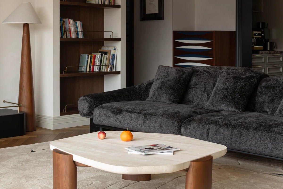 A modern living room featuring a dark gray textured sofa, a low organic-shaped coffee table with a light stone top and wooden legs, a large white sculptural pendant light, and built-in wooden bookshelves.