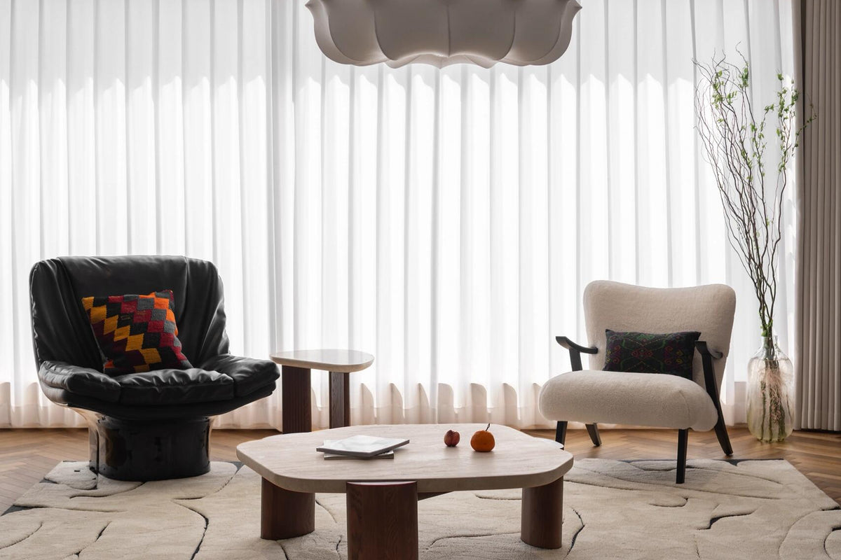 A modern living room interior featuring a sculptural white pendant light hanging from a decorative ceiling medallion, with a low wooden coffee table, a black leather armchair with a colorful pillow, and a cream upholstered armchair in front of sheer white curtains.