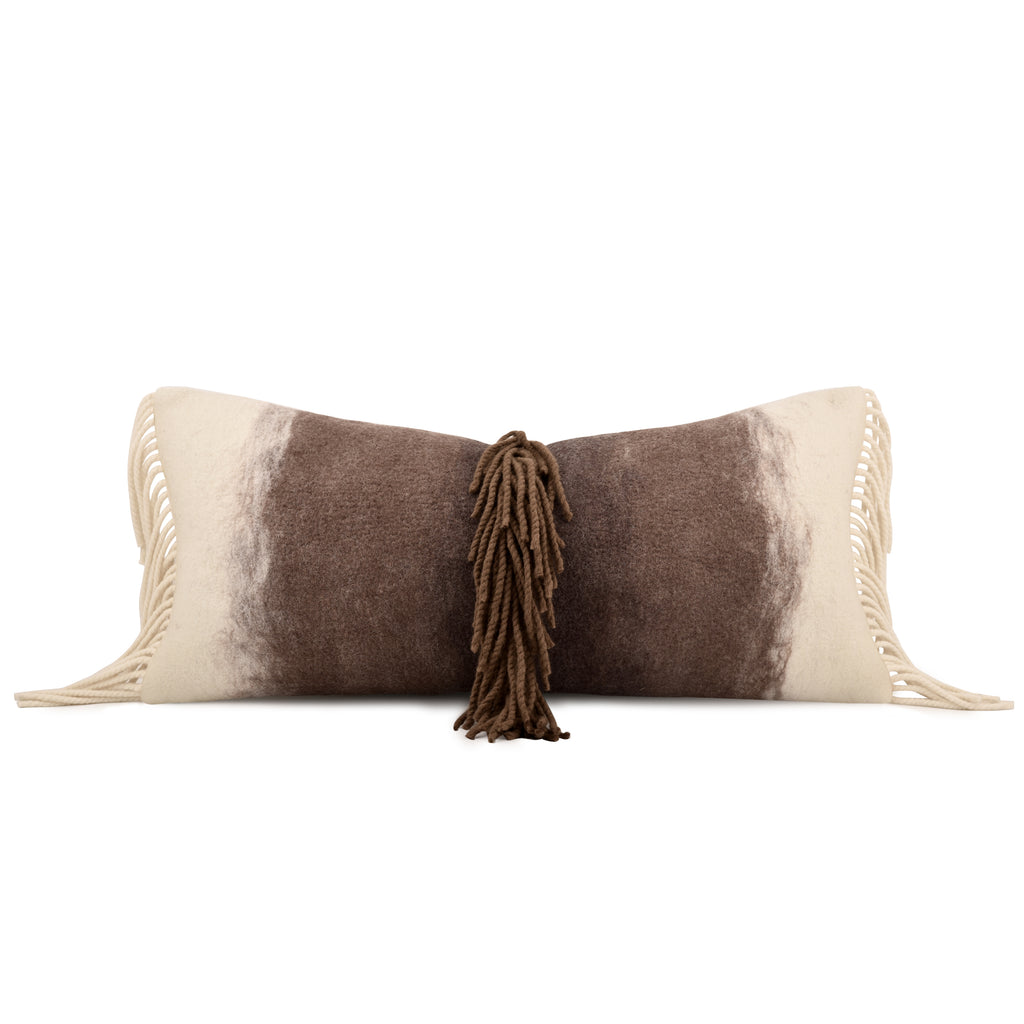 A rectangular throw pillow with a brown and cream cowhide-inspired pattern, featuring cream fringe on the sides and a central brown tassel, shown on a plain white background.