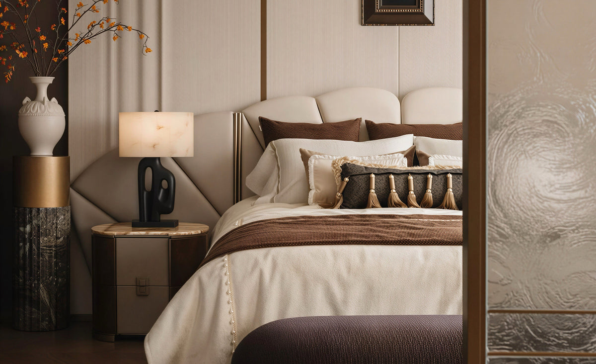 A luxury bedroom interior featuring a bed with cream and brown bedding, a matching tufted headboard, a wooden nightstand with a lit table lamp, and decorative side tables with a white vase and dried branches.